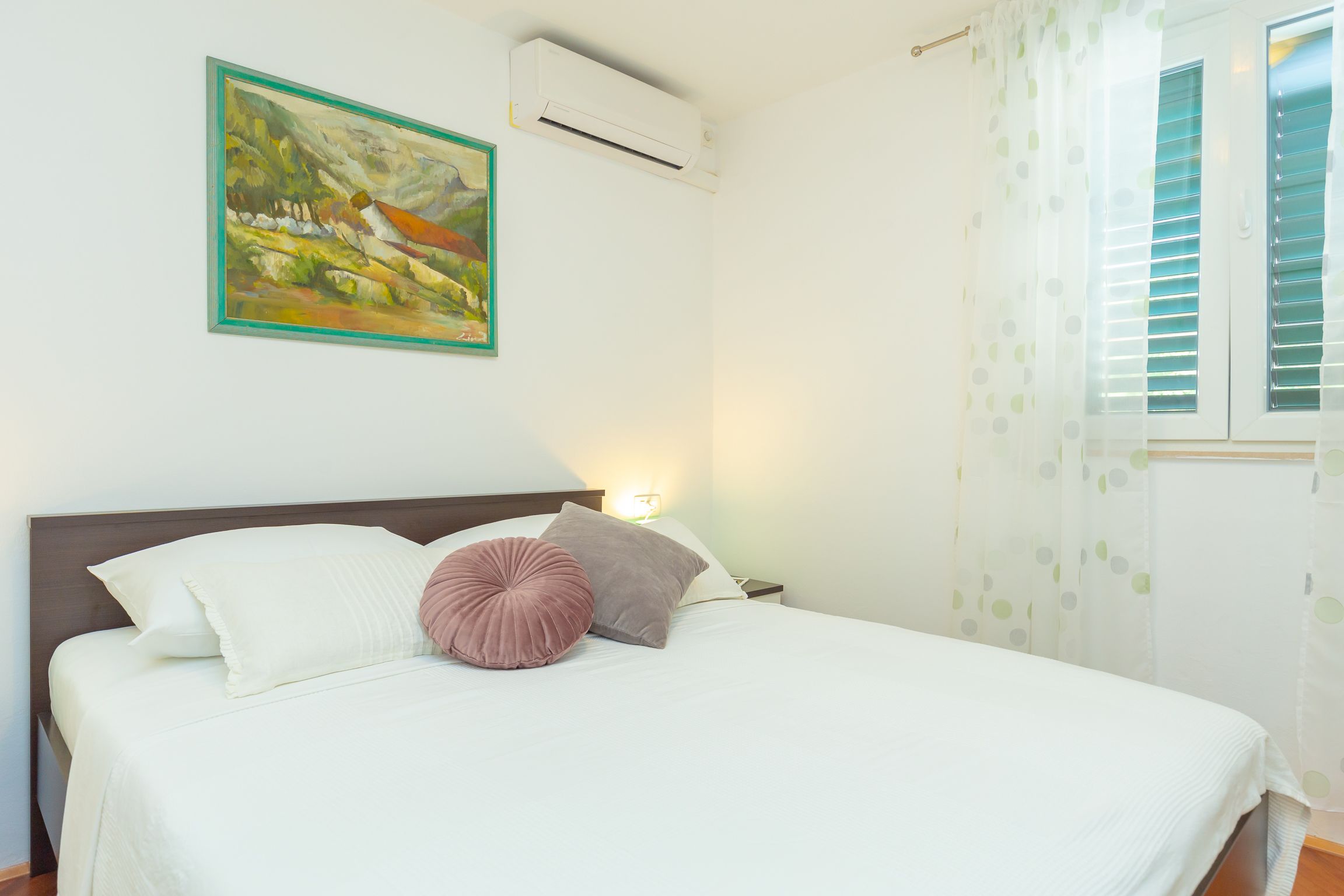 A neatly made bed with white and pink pillows, a painting on the wall, and a window with blinds.