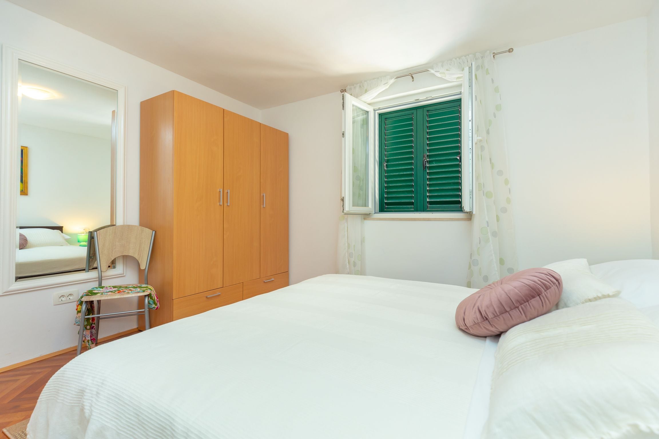 A bedroom with a bed, a wooden wardrobe, a window with green shutters, and a chair with a colorful cushion.