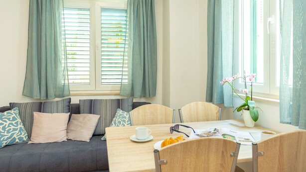 A cozy room with a sofa, a dining table with chairs, a potted plant, and a window with curtains. - Apartment Colette (Dubrovnik)