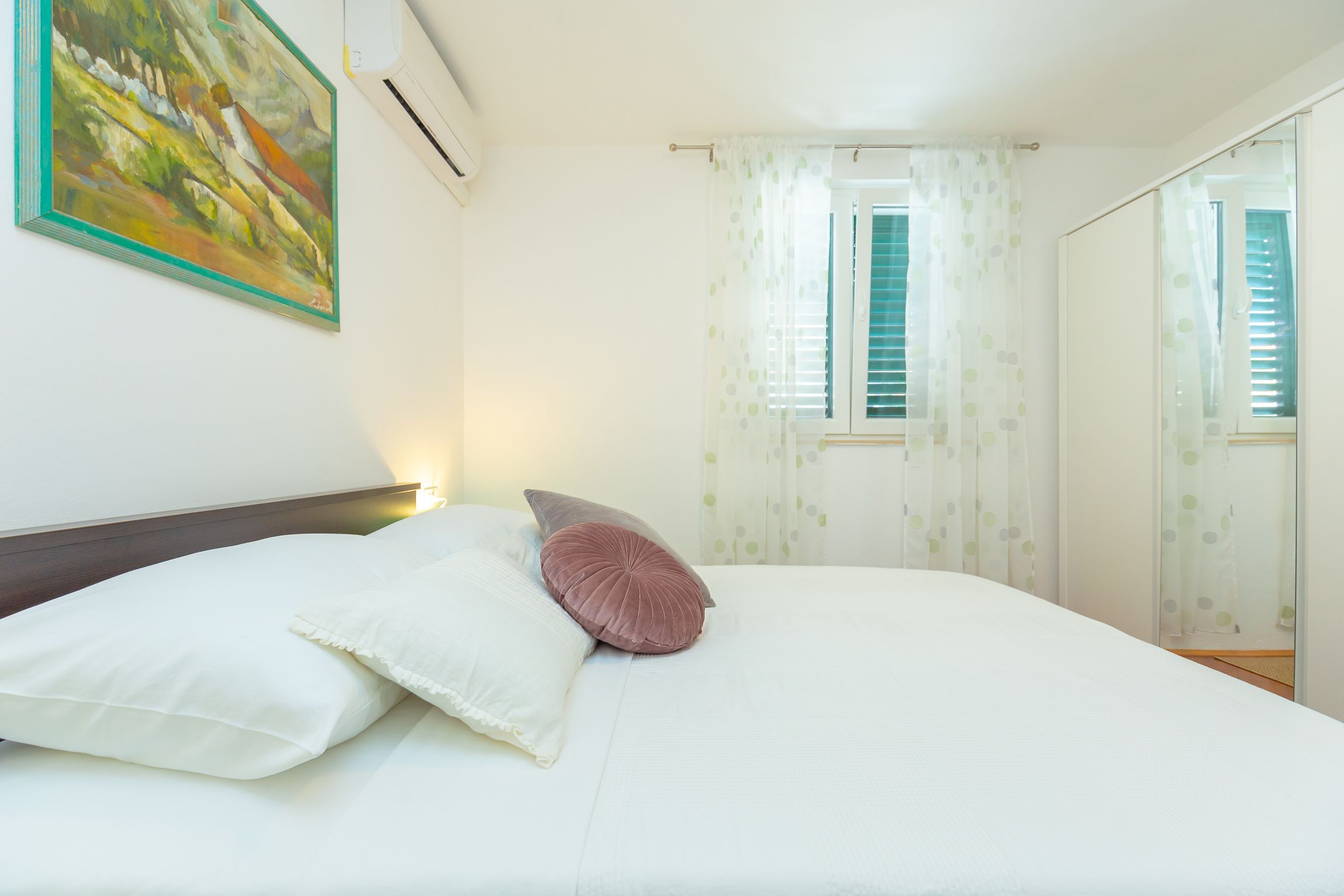 A neatly made bed with white and pink pillows, a painting on the wall, and a window with curtains.