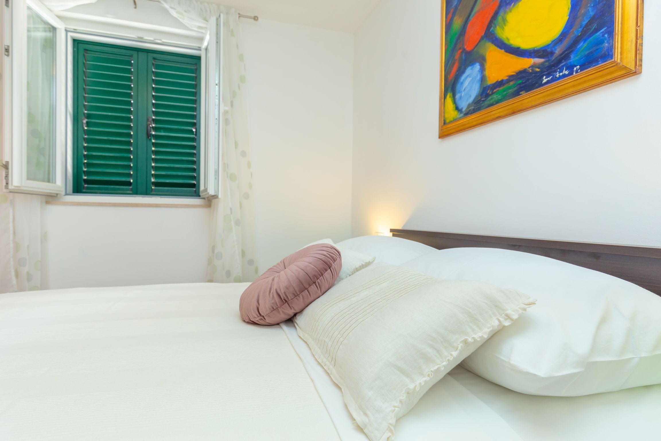 A bedroom with a bed, pillows, a window with shutters, and a painting on the wall.