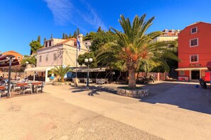 Experience the charm of a Mediterranean town's central square, surrounded by traditional and modern architecture, with a central palm tree and outdoor seating areas.