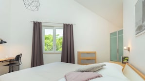 Ein Schlafzimmer mit Bett, Schreibtisch, Stuhl, einem Fenster mit Vorhängen, einem Kleiderschrank und einer Wandleuchte.