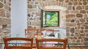 A stone-walled room with a glass table, chairs, a plate with food, and a window with a view of greenery. - Hedera Estate, Hedera A20 Apartment (Dubrovnik)