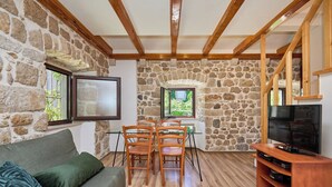 A room with a stone wall, wooden beams, a sofa, a dining table with chairs, a TV, and a window. - Hedera Estate, Hedera A20 Apartment (Dubrovnik)