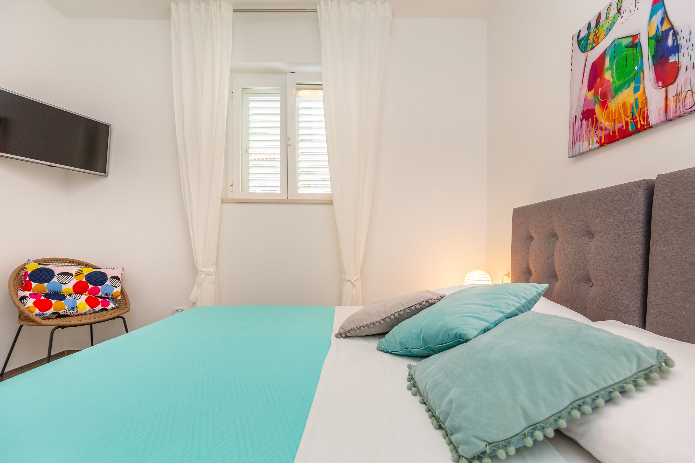 A bedroom with a bed, a television, a chair with a colorful cushion, and a painting on the wall.