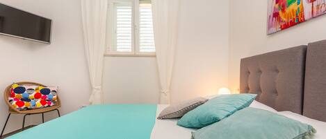A bedroom with a bed, a television, a chair with a colorful cushion, and a painting on the wall.