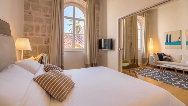 A hotel room with a bed, a TV, a sofa, and a view of palm trees through a window. - Hedera Estate, Hedera A4 (Dubrovnik)