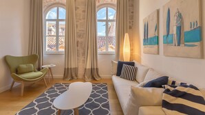 A room with a sofa, a chair, a small table, and a patterned rug. There are two windows with curtains, and artwork on the wall. - Hedera Estate, Hedera A4 (Dubrovnik)