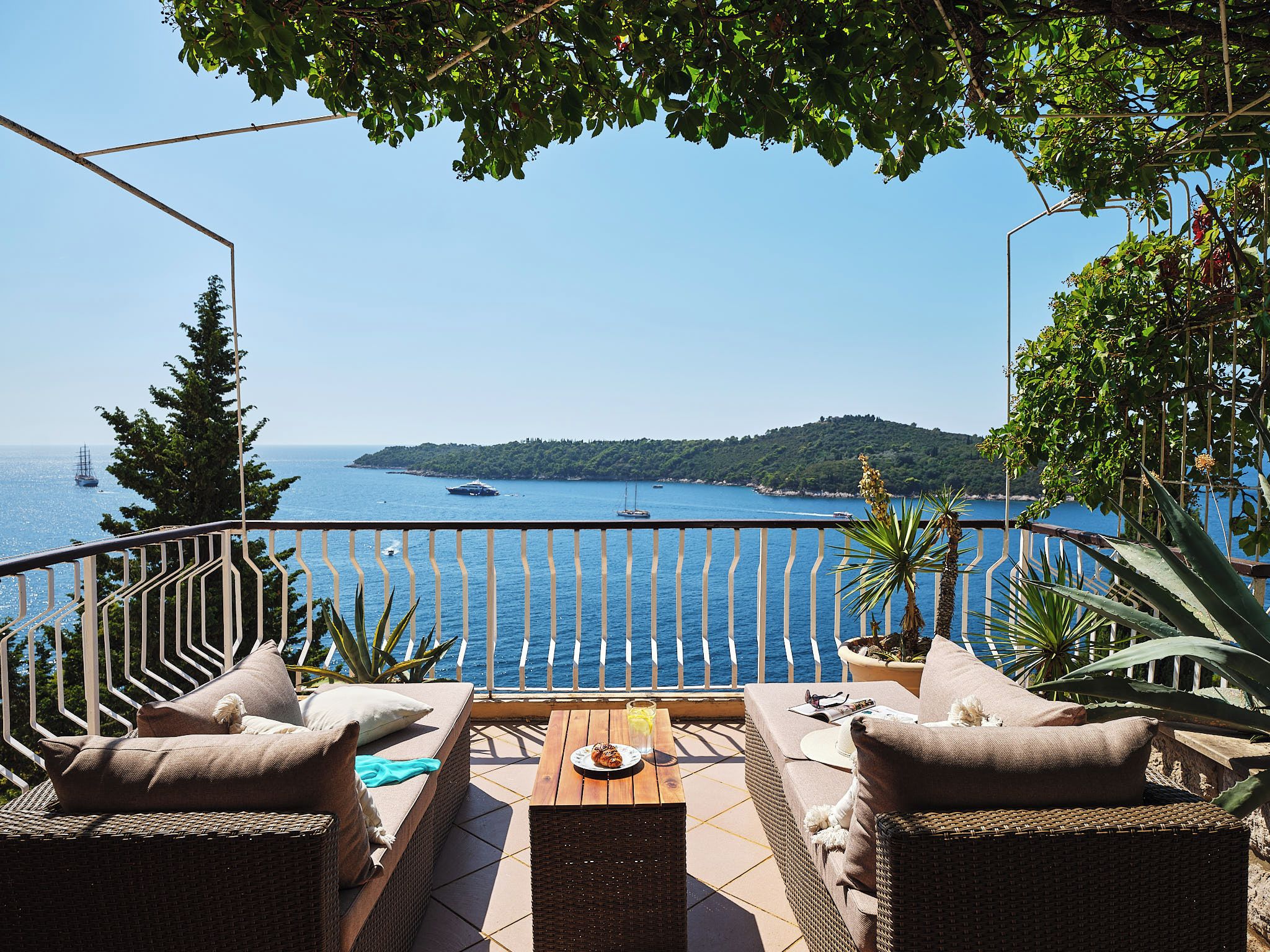 A terrace with wicker furniture, a table, and a view of the sea and a distant island.