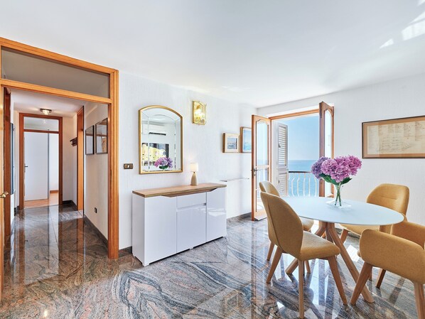 A modern hotel room with a marble floor, a dining area with a white table and yellow chairs, a large mirror, and a view of the sea through a window.