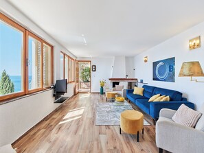 A spacious living area with a blue sofa, a flat-screen TV, and a view of the sea through large windows.
