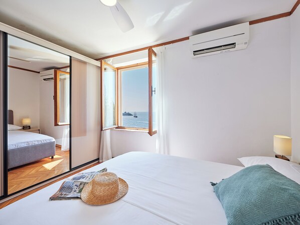 A hotel room with a bed, a window with a view of the sea, a wall-mounted air conditioner, and a bedside lamp.