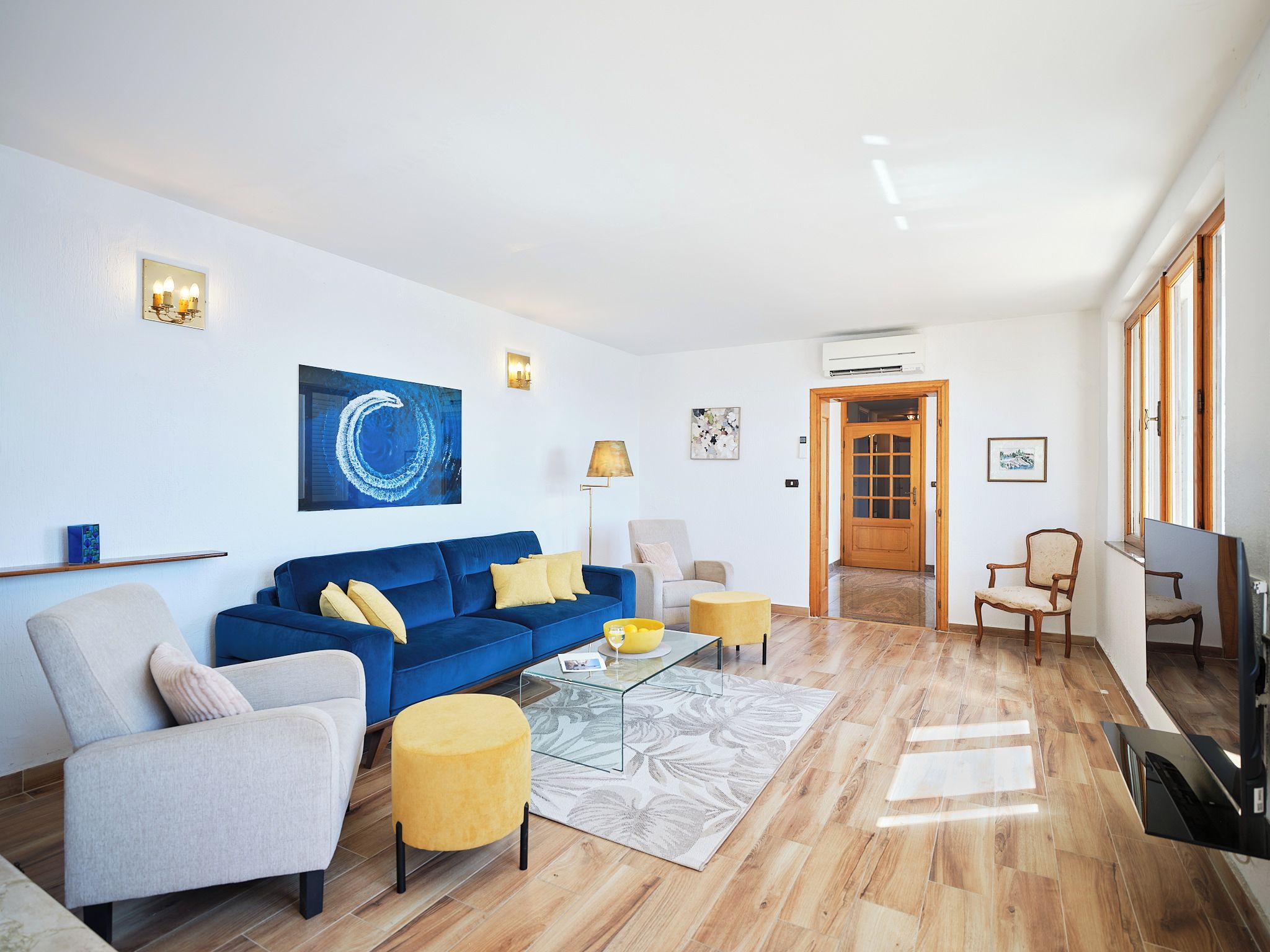 A modern living room with a blue sofa, a grey armchair, and a yellow ottoman. There is a glass coffee table with a bowl and a wine glass. The room has wooden flooring and white walls.