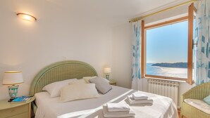 A neatly made bed with pillows and folded towels, a bedside lamp, a wicker chair, and a window with a view of the sea. - Hedera Estate, Hedera A17 (Dubrovnik)