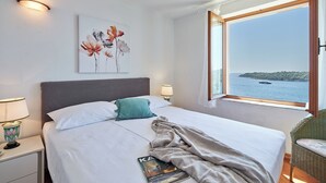 A hotel room with a bed, a nightstand, a lamp, a window with a view of the sea, and a painting on the wall. - Hedera Estate, Hedera A17 (Dubrovnik)