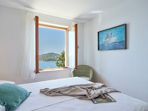 A bedroom with a bed, a window with curtains, a chair, a painting, and a view of a body of water and hills. - Hedera Estate, Hedera A17 (Dubrovnik)