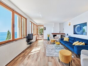 A spacious living area with a large window overlooking the sea, a blue sofa, and a flat-screen TV.