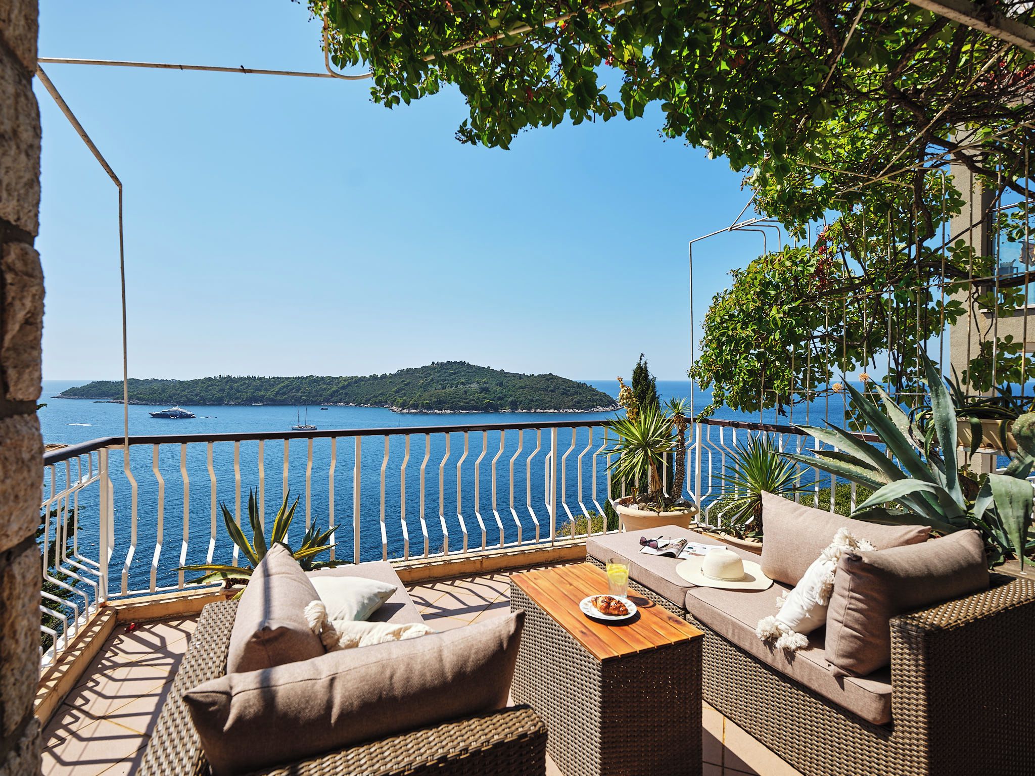 A terrace with wicker furniture, a table, and a view of the sea and a distant island.