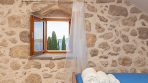 A stone-walled room with a bed, a window with a view of trees and a body of water, and a curtain. - Summer Cottage (Dubrovnik)