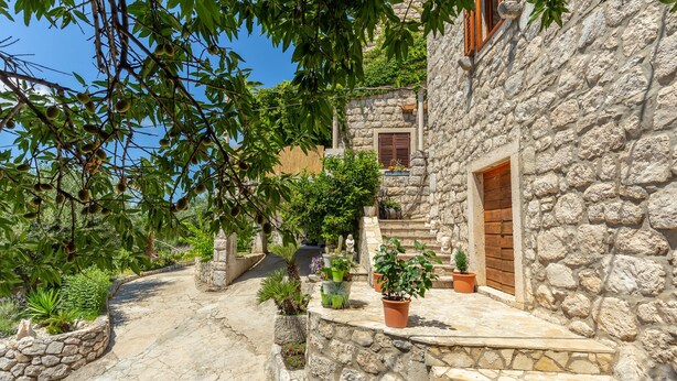 A stone pathway leading to a rustic stone building with a wooden door, surrounded by potted plants and greenery. - Summer Cottage (Dubrovnik)