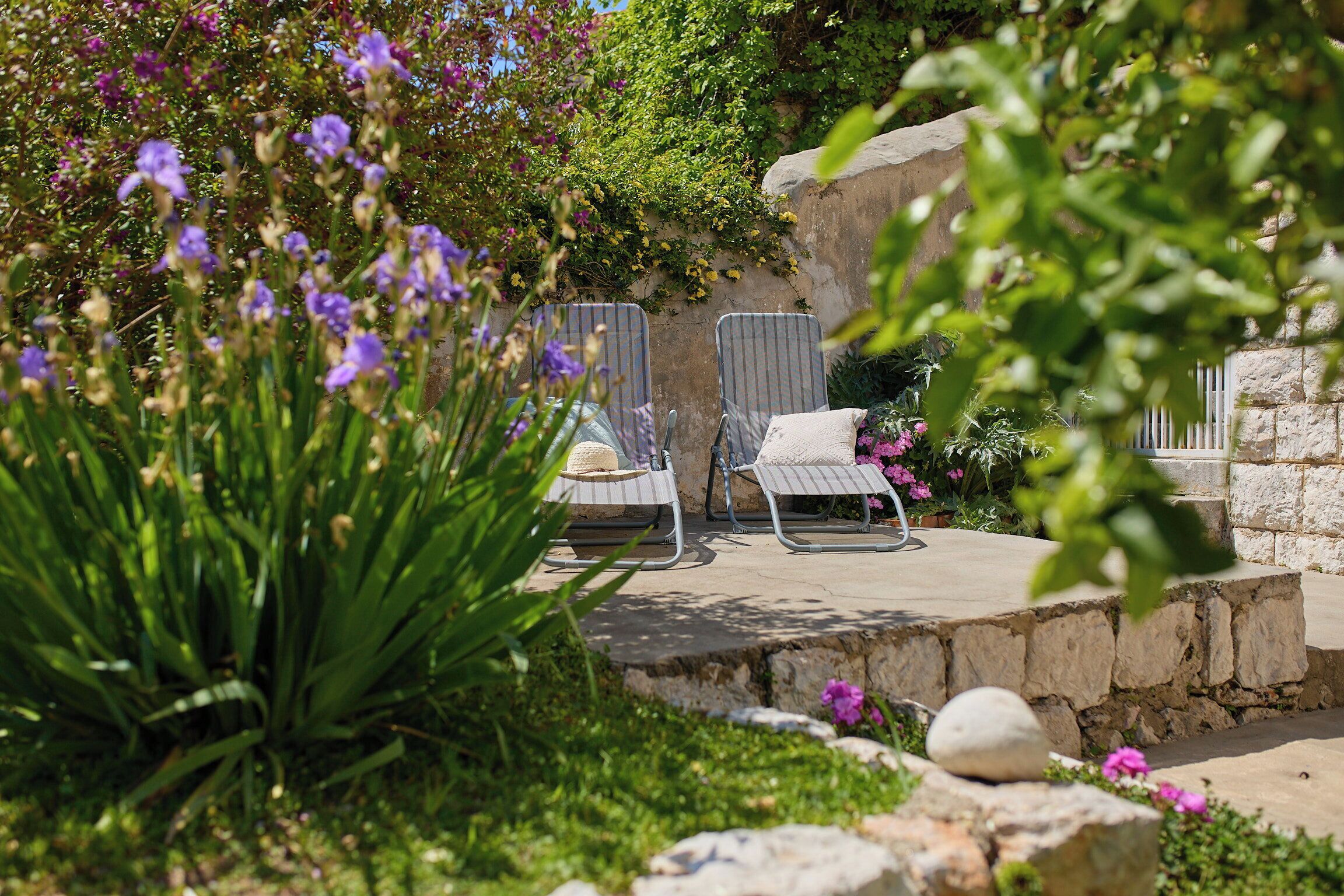 A garden patio with two lounge chairs, a stone wall, and lush greenery.