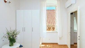 A room with a white wardrobe, a small table with a vase, a window with a view of a tiled roof, and an air conditioning unit. - Hedera Estate, Hedera A34 (Dubrovnik)