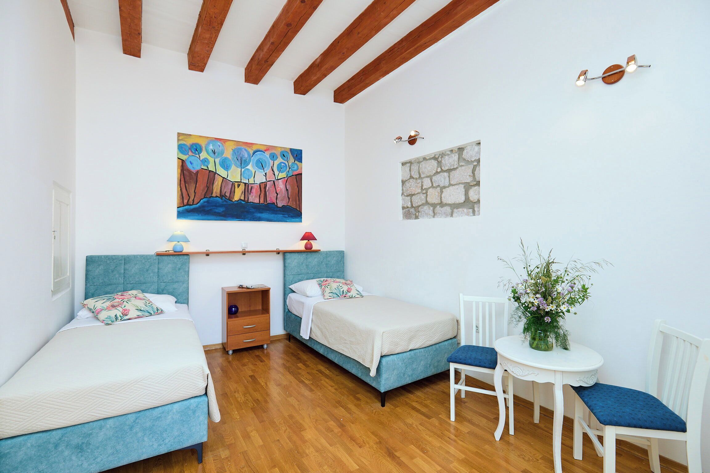 A bedroom with two beds, a small table with a plant, a painting on the wall, and a stone feature.