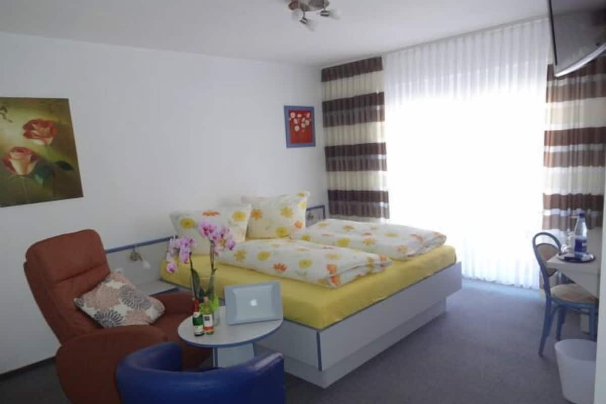 A hotel room with a bed, a chair, a small table, and a window with curtains.