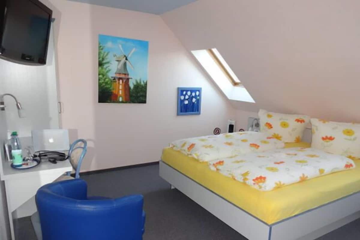 A bedroom with a bed, a desk, a chair, a painting of a windmill, and a skylight.