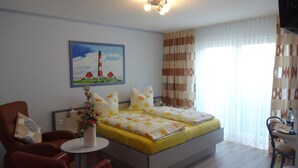 Habitación de hotel con cama, una silla, una mesita y un cuadro de un faro, además de cortinas.