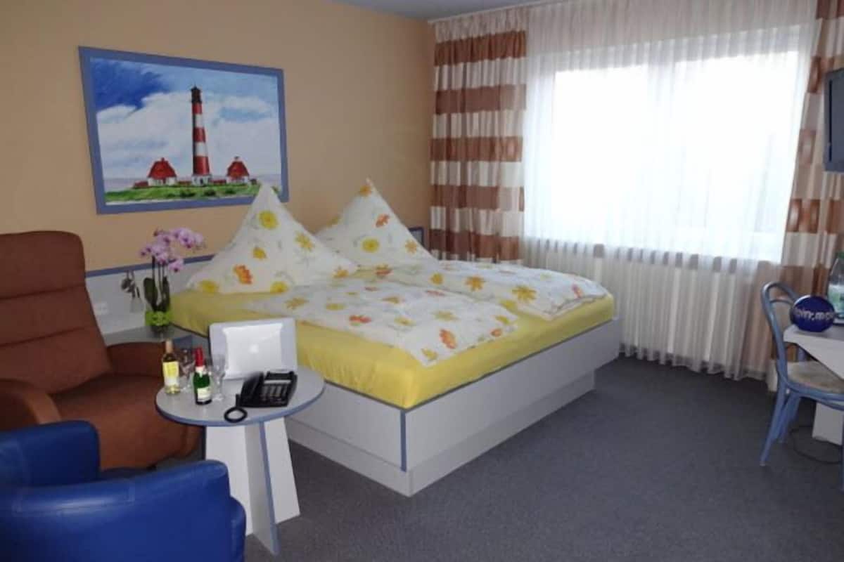 A hotel room with a bed, a desk, a chair, a small table with a phone and bottles, a painting of a lighthouse, and curtains.