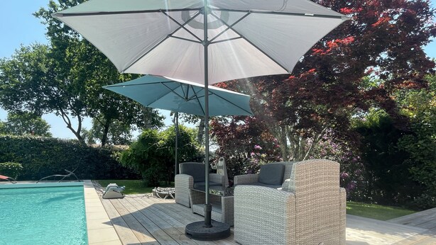 A poolside area with a wicker sofa and umbrella, surrounded by a wooden deck and a pool.