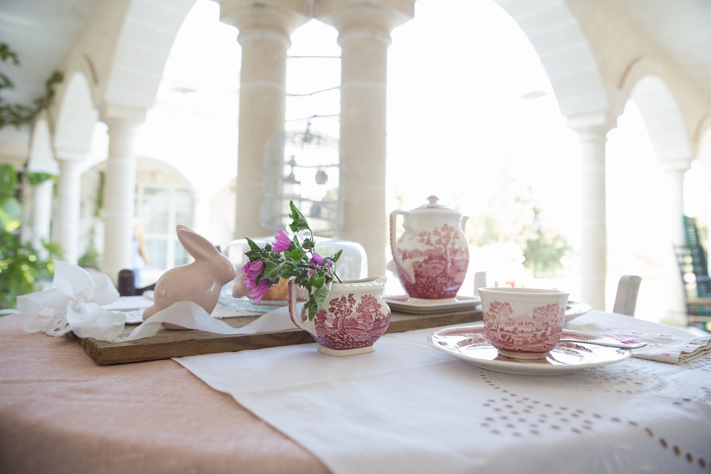 Una mesa dispuesta con vajilla de té en rosa y blanco, un conejo de cerámica y un florero con flores, todo bajo un arco blanco.