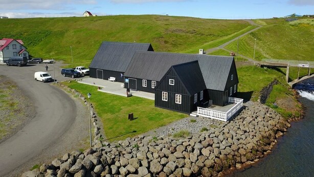 A black building with a white porch, situated by a rocky shoreline with a clear view of a river. - Frændgardur (Skagafjörour)