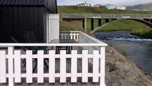 A wooden dock with a white railing, a black building, and a stone bridge over a river. - Frændgardur (Skagafjörour)