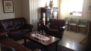 A living room with a dark brown leather sofa, a wooden coffee table with a lace doily, a glass cabinet, and a window with curtains. - Frændgardur (Skagafjörour)