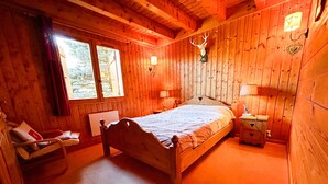 A wooden cabin bedroom with a bed, a chair, a nightstand, a lamp, and a deer head mounted on the wall. - Chalet for up to 16 guests in Bernex, pets welcome (Bernex)