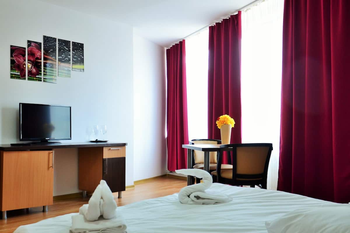 A hotel room with a bed, a desk with a TV, a chair, a small table with a vase, and curtains.