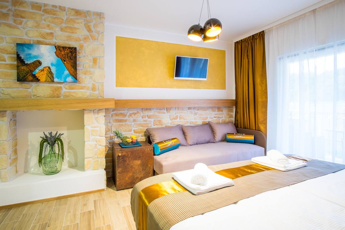 A hotel room with a stone wall, a flat-screen TV, a bed with a gold bedspread, a wooden nightstand, and a vase with plants.
