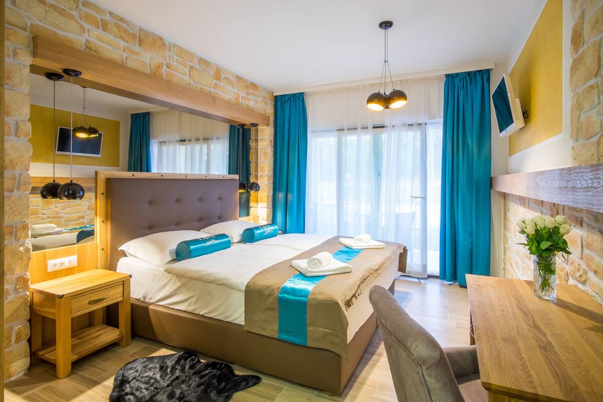 A hotel room with a stone wall, a bed with blue accents, a wooden desk, and a mirror.