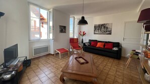 A living room with a wooden coffee table, a black sofa with red cushions, a red chair, a TV on a stand, and a large window with a view of buildings.