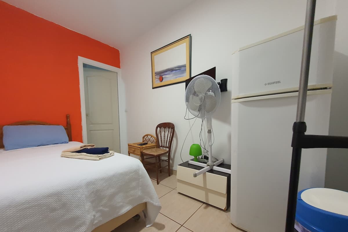 A bedroom with a bed, a chair, a standing fan, a heater, and a white wardrobe.