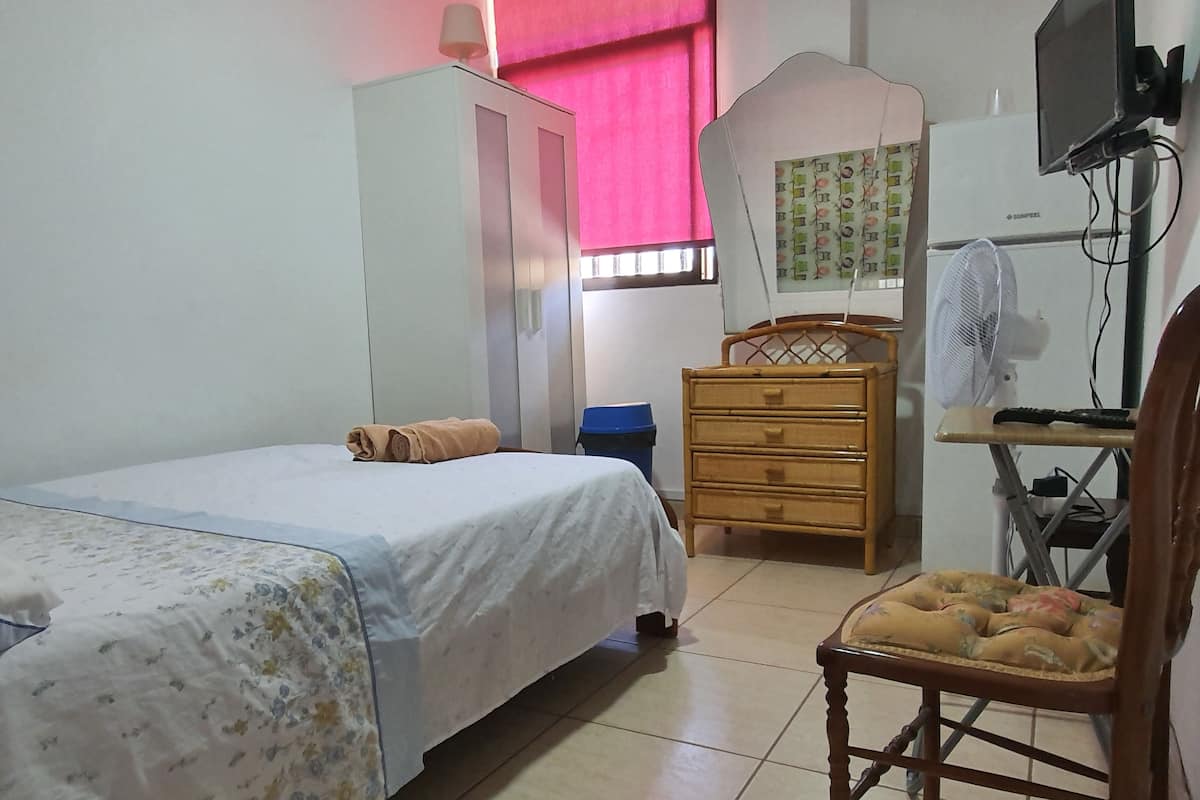 A bedroom with a bed, a wooden dresser, a chair, a desk with a computer, a TV, and a fan.