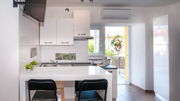 A modern kitchen with white cabinetry, a central island with two black chairs, and a view of a balcony with a plant. - Apartments Karolina (Opatija)