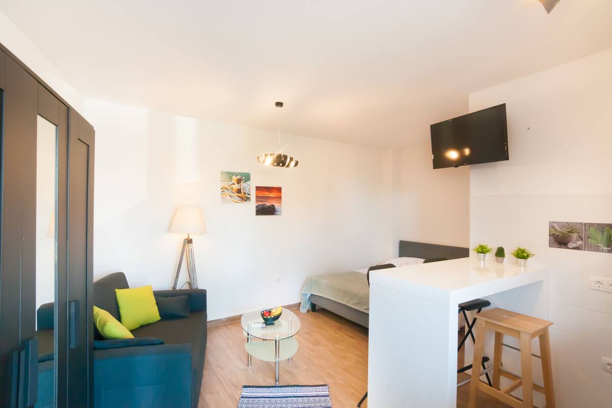 Un espacio moderno para vivir con sofá, una mesita redonda, cama y una pequeña cocina.