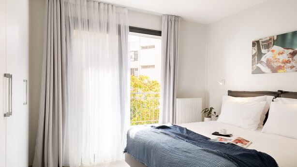 A modern hotel room with a large bed, a window with sheer curtains, a view of greenery outside, and a painting on the wall. - Thess Residences (Thessaloniki)