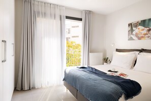 Ett modernt hotellrum med en stor säng, ett fönster med tunna, genomskinliga gardiner, utsikt över grönområden och en tavla på väggen.