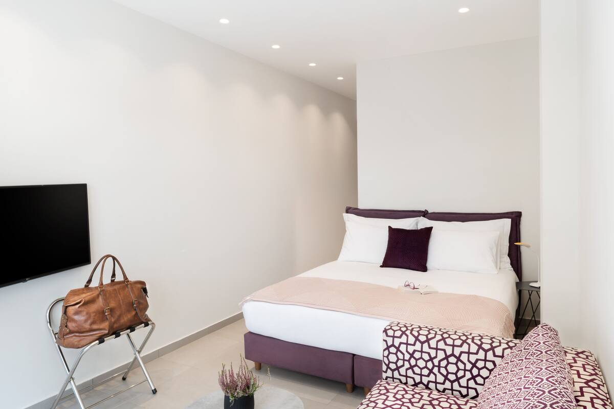 A modern bedroom with a bed, a TV mounted on the wall, a brown leather bag on a chair, and a small potted plant.