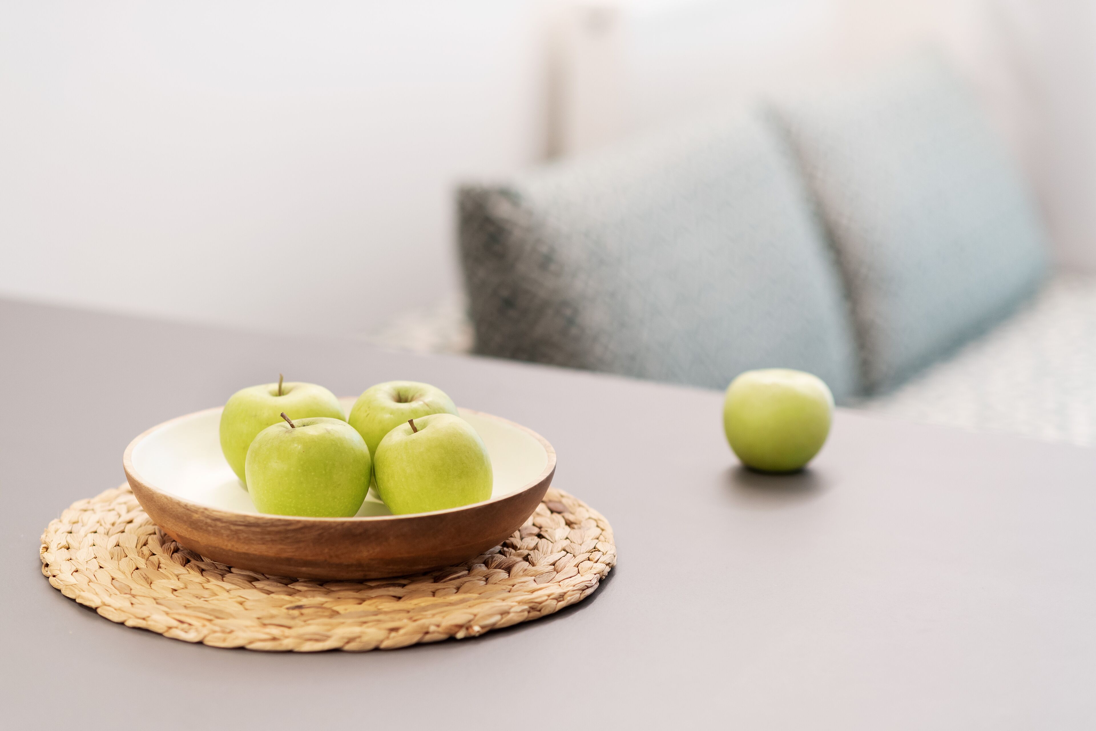 A bowl of green apples on a woven placemat on a table.